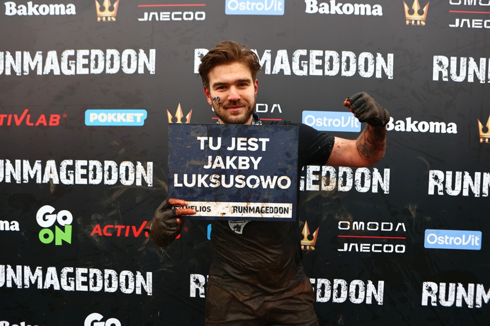 Szymon Mojsak po biegu Runmageddon — ubłocony, trzyma tabliczkę organizatorów z napisem TU JEST JAKBY LUKSUSOWO